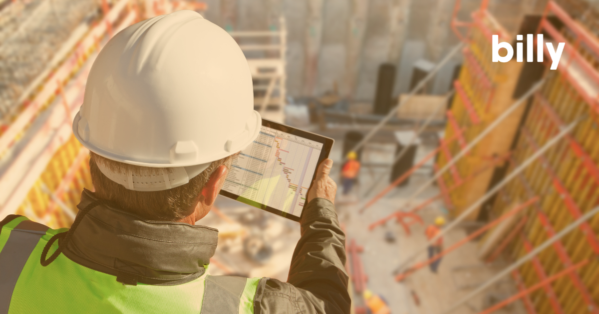 Man in a hardhat on a building during construction holding a tablet while looking at a document.